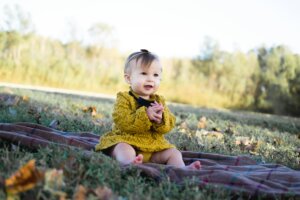 Cute baby girl sitting on a blanket in a park during fall, smiling happily.