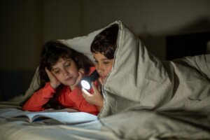 Two children reading a book under a blanket with a flashlight at night, sharing a cozy bedtime story.