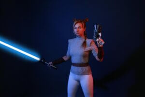 A woman dressed in sci-fi costume wields a lightsaber and blaster in a dramatic studio setting.